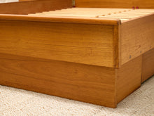 Load image into Gallery viewer, Close-up of a wooden bed frame with a warm honey finish, showing the slatted headboard and side panel resting on a textured cream carpet.
