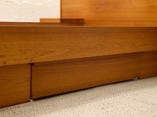Load image into Gallery viewer, Close-up of a mid-century modern wooden bed frame with a warm brown finish, showing the side panel, footboard, and storage drawers on a textured beige carpet.

