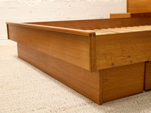 Load image into Gallery viewer, A close-up of a mid-century modern wooden platform bed frame with a warm medium-brown finish, slatted headboard, and storage drawers underneath, set on a textured beige carpet against a white brick wall.
