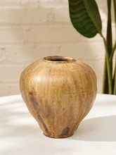 Load image into Gallery viewer, A rustic ceramic vase with a rounded bulbous shape and a small flared opening, shown on a white surface against a light brick wall with a green plant leaf in the background.
