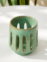 Load image into Gallery viewer, A small ceramic candle holder with a light green glazed finish and a rustic brown rim, featuring vertical oval cutouts, set on a white textured surface.
