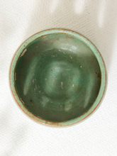 Load image into Gallery viewer, A small round ceramic bowl with a glossy green glaze and a natural terracotta-colored rim, photographed from directly above on a white textured fabric background.

