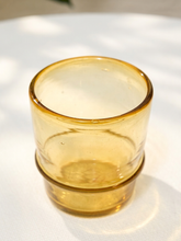 Load image into Gallery viewer, A small amber yellow translucent glass tumbler with a thick base, resting on a white textured surface with soft natural shadows.
