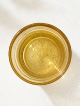 Load image into Gallery viewer, A top-down view of a small, round amber glass cup or dish on a textured white fabric background.
