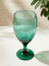 Load image into Gallery viewer, A translucent green glass goblet with a wide bowl, stem, and circular base, set on a white textured fabric surface with soft shadows and a blurred leafy plant in the background.
