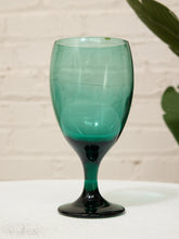Load image into Gallery viewer, A translucent teal green wine glass with a dark green base, standing on a white textured surface against a white brick wall background with a green leaf in the upper right corner.
