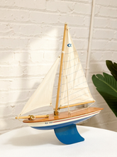 Load image into Gallery viewer, A wooden model sailboat with cream-colored sails, a light wood hull, and a blue base, displayed on a white surface against a white brick wall background.
