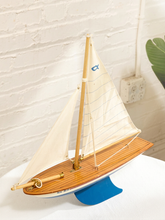 Load image into Gallery viewer, A wooden model sailboat with cream-colored sails, a brown wood-grain deck, and a blue hull and support stand, displayed on a white surface against a white brick wall.
