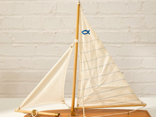 Load image into Gallery viewer, A wooden model sailboat with cream-colored canvas sails, blue stitched lines, and a small blue fish emblem on the sail, displayed on a wooden base against a white brick wall background.
