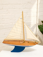 Load image into Gallery viewer, A wooden model sailboat with cream-colored sails, a blue hull, and a blue wave-shaped base, displayed on a white surface against a white brick wall.
