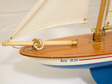 Load image into Gallery viewer, Close-up of a decorative wooden model sailboat with cream canvas sails, a warm brown wooden deck, brass fittings, and a blue hull section. The side of the boat reads 'Acts 20:24' in blue text.
