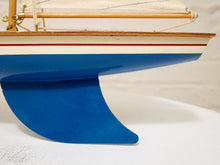 Load image into Gallery viewer, Close-up of a model sailboat hull with a bright blue bottom, white upper section, thin red stripe, and natural wood trim and mast against a light neutral background.

