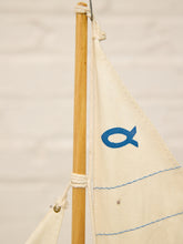 Load image into Gallery viewer, Close-up of a small sailboat with a light wood mast and an off-white canvas sail featuring a blue fish symbol and blue stitched lines, set against a light neutral background.
