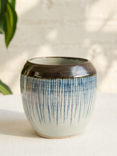 Load image into Gallery viewer, A handmade ceramic pot with a glossy dark brown rim and a body featuring vertical blue brushstroke patterns on a white base, set on a textured white surface with a soft-focus background.
