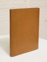 Load image into Gallery viewer, A tan textured leather folio or notebook cover standing upright on a white surface against a white brick wall background.
