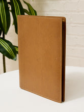 Load image into Gallery viewer, A tan textured leather folio or notebook cover standing upright on a white surface, with a white brick wall and green plant leaves in the background.
