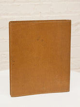 Load image into Gallery viewer, A tan textured faux-leather binder or folder standing upright against a white brick wall background.
