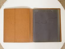 Load image into Gallery viewer, An open tan leather portfolio folder or binder with clear plastic sleeves on the right side, photographed from above on a white round surface.
