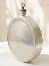 Load image into Gallery viewer, A vintage oval-shaped silver metal canteen with a small hanging loop on top, displayed on a white surface against a white brick wall with soft shadows.
