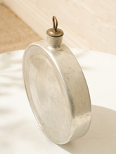 Load image into Gallery viewer, A vintage oval metal canteen or flask with a small hanging loop on a brass cap, resting on a white textured surface with soft shadows.

