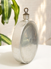 Load image into Gallery viewer, A vintage oval-shaped silver metal flask with a top loop handle, standing on a white surface with a white brick wall and green plant leaves in the background.

