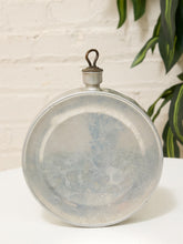 Load image into Gallery viewer, A vintage oval-shaped translucent glass bottle with a metal screw cap and hanging loop, displayed on a white surface against a white brick wall with green leaves in the background.
