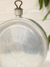 Load image into Gallery viewer, A vintage round glass flask with a metal screw cap and loop handle, showing a faded embossed logo on the bottom, set against a light brick wall background with green leaves.
