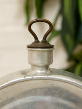 Load image into Gallery viewer, Close-up of a vintage silver metal pot lid featuring a dark brown wooden loop handle on a metal cap, set against a soft-focus background of green leaves and a light wall.
