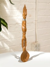 Load image into Gallery viewer, A tall, hand-carved wooden spoon with a stylized human figure handle and a carved face at the bowl, standing upright on a white surface against a white brick wall.

