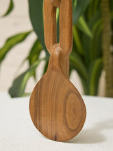 Load image into Gallery viewer, A close-up of a hand-carved wooden spoon with a unique loop handle, resting on a white textured surface with a blurred green plant in the background.
