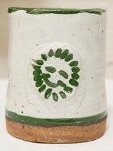 Load image into Gallery viewer, A handmade ceramic cup with a textured off-white glaze, featuring a green painted circular leaf pattern on the front and a natural brown clay base.
