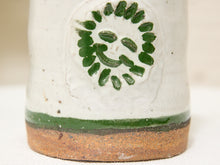 Load image into Gallery viewer, Close-up of a handmade ceramic pot or vase showing a textured off-white glaze, a painted green smiley face motif, a green horizontal band, and a rustic brown terracotta foot ring.
