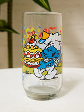 Load image into Gallery viewer, A clear glass drinking tumbler featuring a colorful Smurfs cartoon illustration, including a blue Smurf carrying a yellow cake and a white Smurf wearing a chef's hat. The words 'SMURFS' are printed near the bottom.
