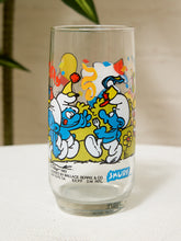 Load image into Gallery viewer, A vintage clear glass Smurf-themed beer tumbler featuring colorful cartoon Smurf characters and the 'SMURF' logo, photographed on a white surface with a soft beige background.
