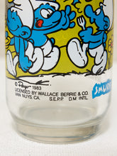Load image into Gallery viewer, Close-up of a clear glass tumbler featuring a colorful cartoon label with blue Smurf characters on a yellow background and white text near the bottom.
