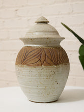 Load image into Gallery viewer, A handmade ceramic lidded jar with a speckled off-white glaze and a textured brown band featuring embossed leaf patterns, set against a white brick wall background.
