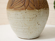 Load image into Gallery viewer, A close-up of a handmade ceramic vase with a textured, speckled off-white body and a brown glazed lid featuring embossed leaf patterns.
