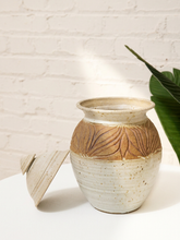 Load image into Gallery viewer, A handmade ceramic jar with a matching lid, featuring a textured beige and brown leaf pattern on the middle section, set against a white brick wall with a green leaf in the background.
