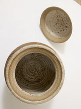 Load image into Gallery viewer, A handmade ceramic jar with a matching lid, shown from a top-down view on a white background. The jar has a rustic, unglazed clay body with a warm beige and brown speckled glaze inside, and a contrasting light beige rim.
