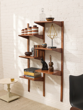 Load image into Gallery viewer, A mid-century modern wooden floating bookshelf with multiple angled shelves, styled with books, decorative vases, a glass decanter, and sculptures against a white brick wall.
