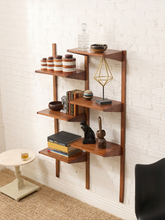 Load image into Gallery viewer, A mid-century modern wooden wall-mounted shelving unit with five tiers, styled with books, decorative vases, and small sculptures against a white brick wall.
