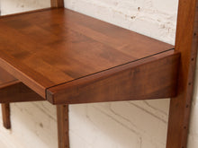 Load image into Gallery viewer, Close-up of a wooden wall-mounted desk shelf with a warm medium-brown finish, showing the tabletop, support bracket, and vertical frame against a white brick wall.
