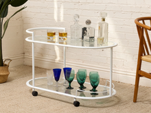 Load image into Gallery viewer, A white metal bar cart with two glass shelves and black caster wheels, styled with various drinking glasses and decanters in a bright room with a white brick wall.
