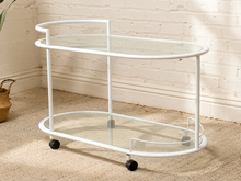 Load image into Gallery viewer, A white metal bar cart with two clear glass shelves and black caster wheels, set against a white brick wall and beige carpet.
