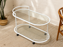 Load image into Gallery viewer, A white metal two-tier bar cart with clear glass shelves and black caster wheels, shown in a styled room corner with a brick wall, carpet, plant, and wooden chair.
