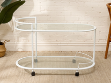 Load image into Gallery viewer, A white metal bar cart with two clear glass shelves and black caster wheels, shown in a styled room setting with a white brick wall, beige rug, and a potted plant.
