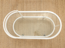 Load image into Gallery viewer, A white metal oval frame with a clear glass insert, photographed from above on a textured beige woven rug.
