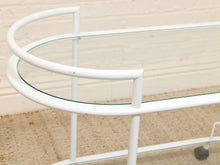 Load image into Gallery viewer, A close-up of a white metal bar cart with a curved glass top, set against a light beige brick wall and carpeted floor.
