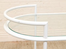 Load image into Gallery viewer, A close-up of a white metal bar cart or side table with a curved glass top and white tubular frame, set against a light beige brick wall background.
