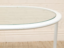 Load image into Gallery viewer, A close-up of a white metal table frame with a clear glass tabletop, set against a light beige brick wall background.
